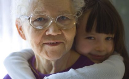 An older woman with white hair and glasses looks at the viewer. A girl of around 5-6 years old has her arms around her neck affectionately. 